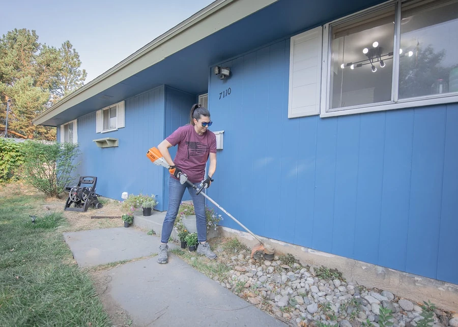 Woman uses a stihl trimmer to cut the grass and weeds that grow up against the house